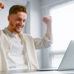 Concentrated businessman signs contract online and raises hands with happiness sitting near white laptop at grey office table