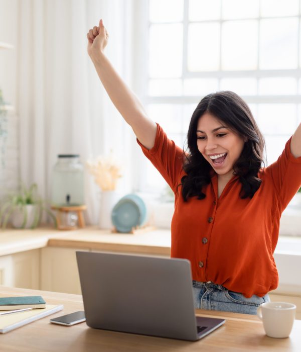 A young woman at a kitchen table expressing excitement with her arms raised and laptop open, working online